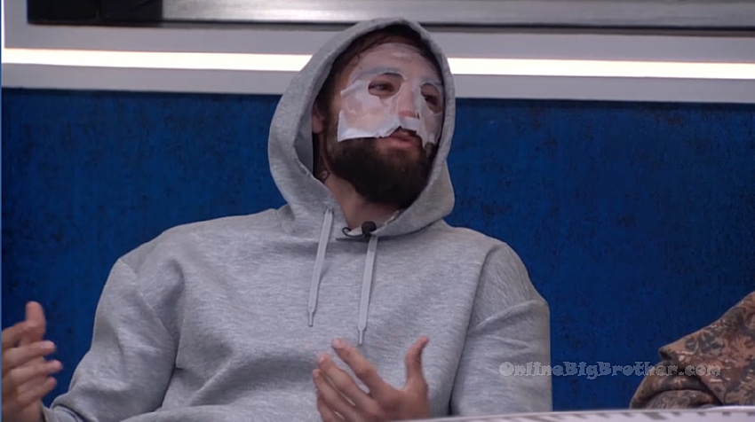 A Big Brother houseguest wearing a sheet face mask and gray hoodie talking with other houseguests after the 8:30 am wake-up call