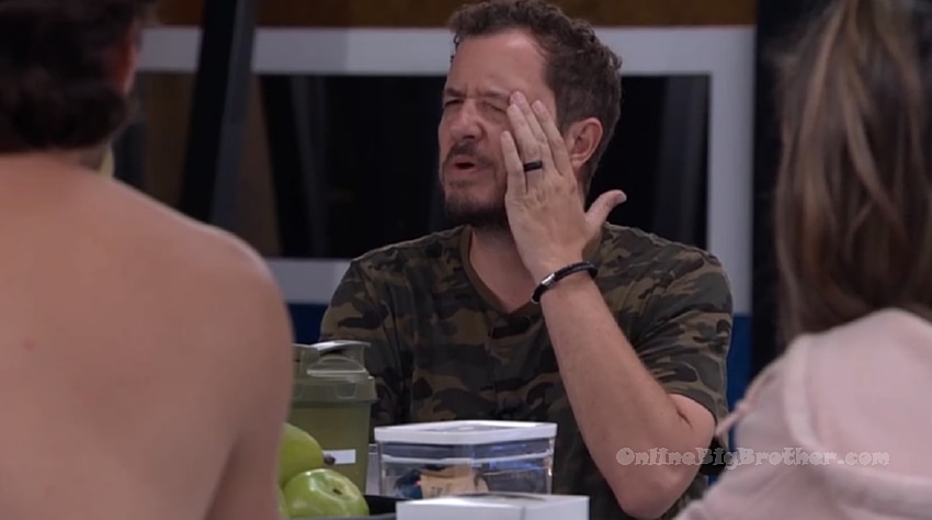 Houseguest in a camo shirt gesturing while talking with others in the kitchen area