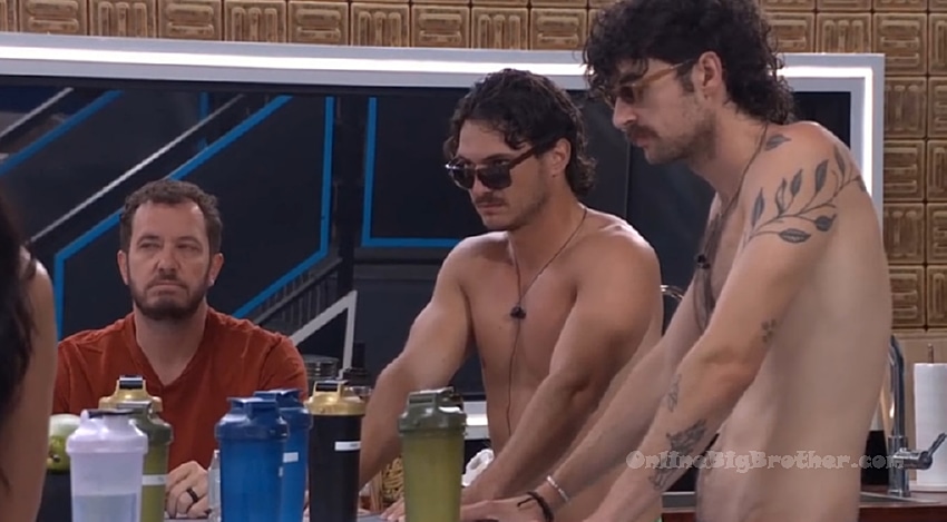Houseguests gathered in the Big Brother kitchen, including two shirtless men and a seated man in a red shirt