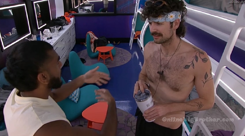 Two Big Brother houseguests talking in the bedroom, one shirtless with a floral headband holding a cup