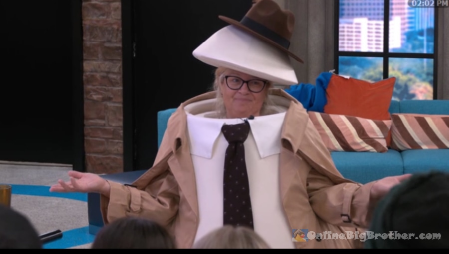 Angela wearing her egg punishment costume — a large egg suit with a hat, collar, tie, and trench coat — while talking to houseguests in the living room