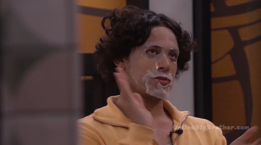 A houseguest wearing a white face mask and yellow shirt talks with others in the Big Brother house at wake up